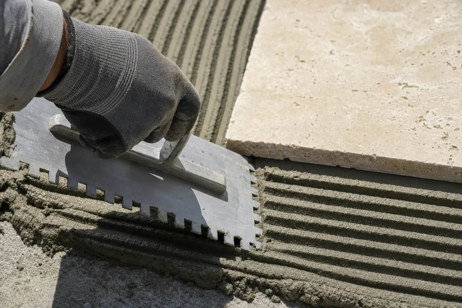 A notched trowel combing ridges into mortar before a travertine paver is set into it